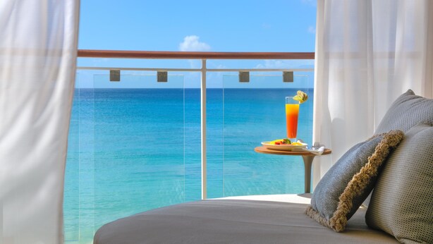 A balcony with a clear view of the ocean, a table with a drink and breakfast, and a bed with pillows. - Fairmont Royal Pavilion (Holetown)