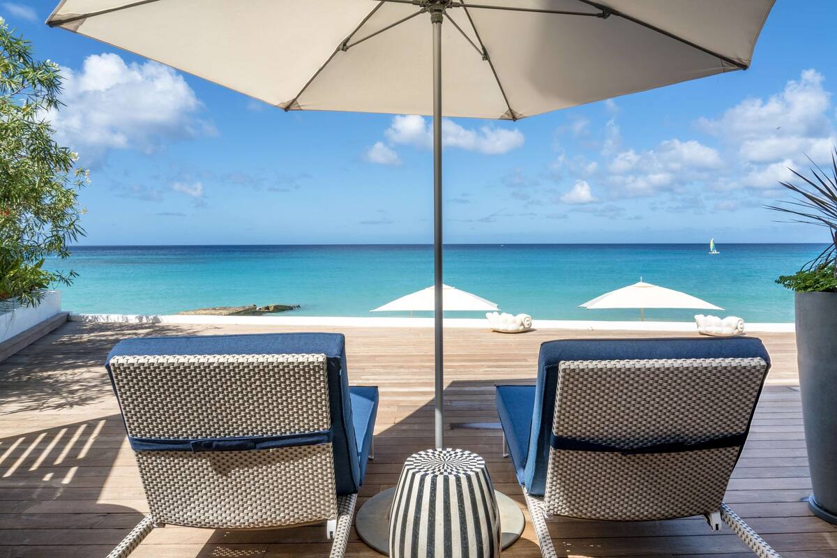 Eine Terrasse mit zwei Korbstühlen, einem gestreiften Hocker und einem großen Sonnenschirm, der Schatten spendet. Die Terrasse bietet Blick auf ein klares, blaues Meer mit einem Segelboot in der Ferne.