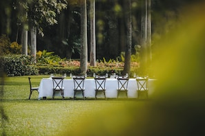 A row of tables set up outdoors with white tablecloths and chairs, surrounded by greenery and trees.