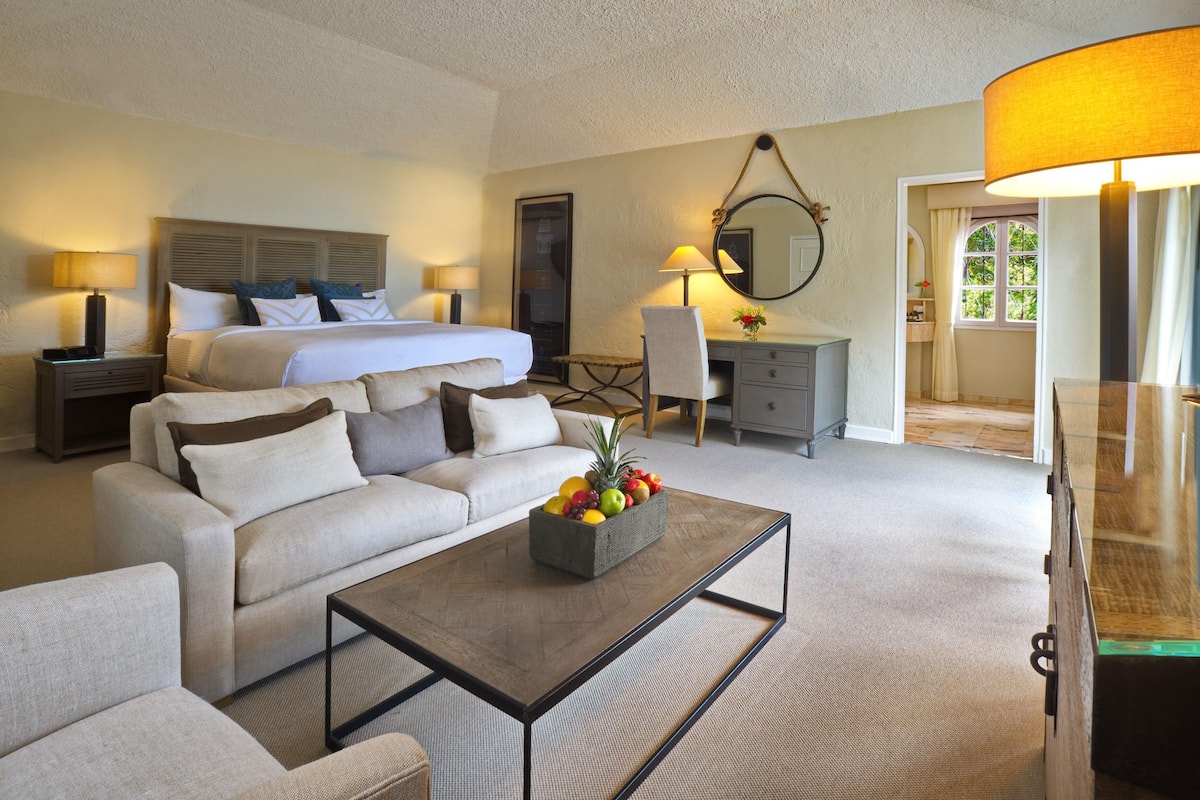 Ein Hotelzimmer mit Bett, Sofa, einem Couchtisch mit Obst, Schreibtisch und Spiegel.