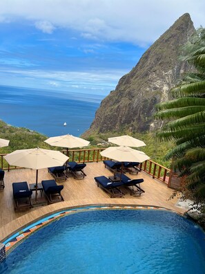 A poolside area with lounge chairs and umbrellas, overlooking a mountainous coastline and the ocean.