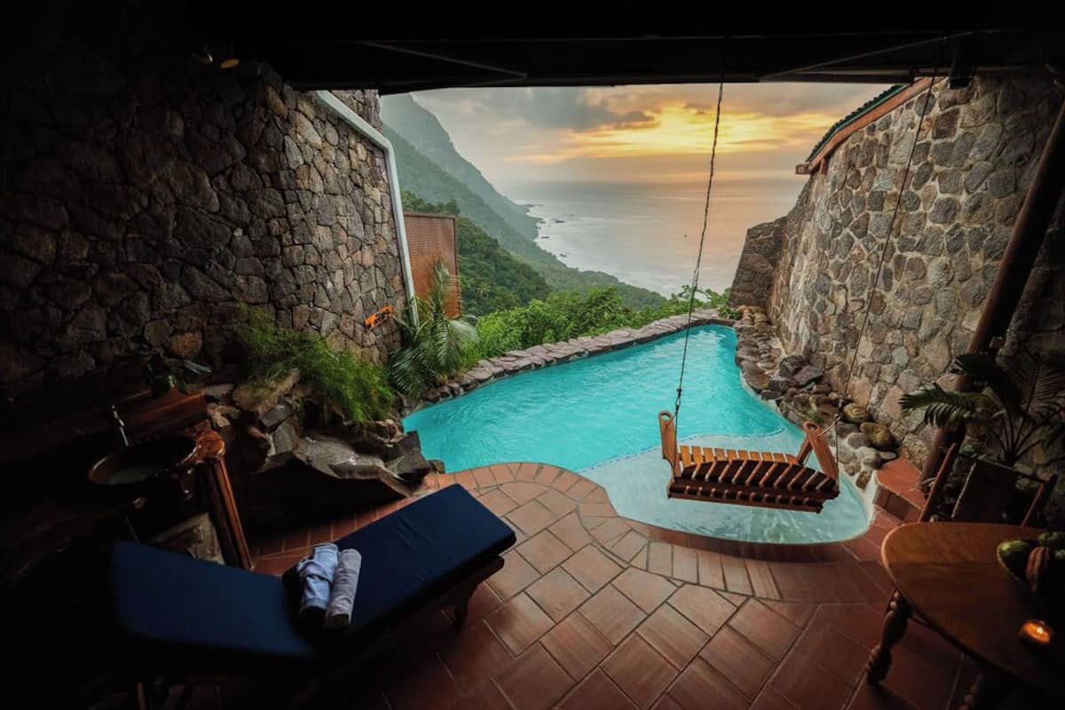 A pool area with a view of the ocean, stone walls, and a wooden swing chair.