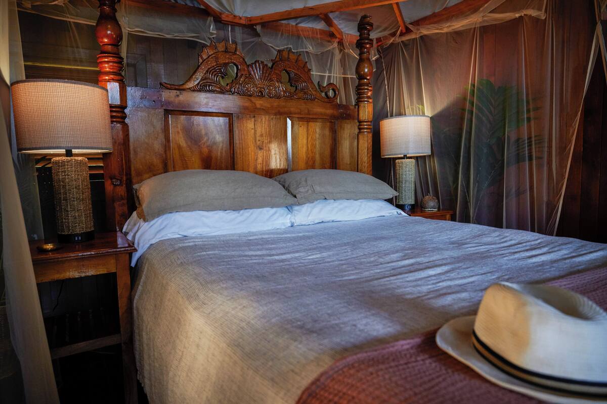 A wooden four-poster bed with a canopy, two bedside lamps, and a hat on the bed.