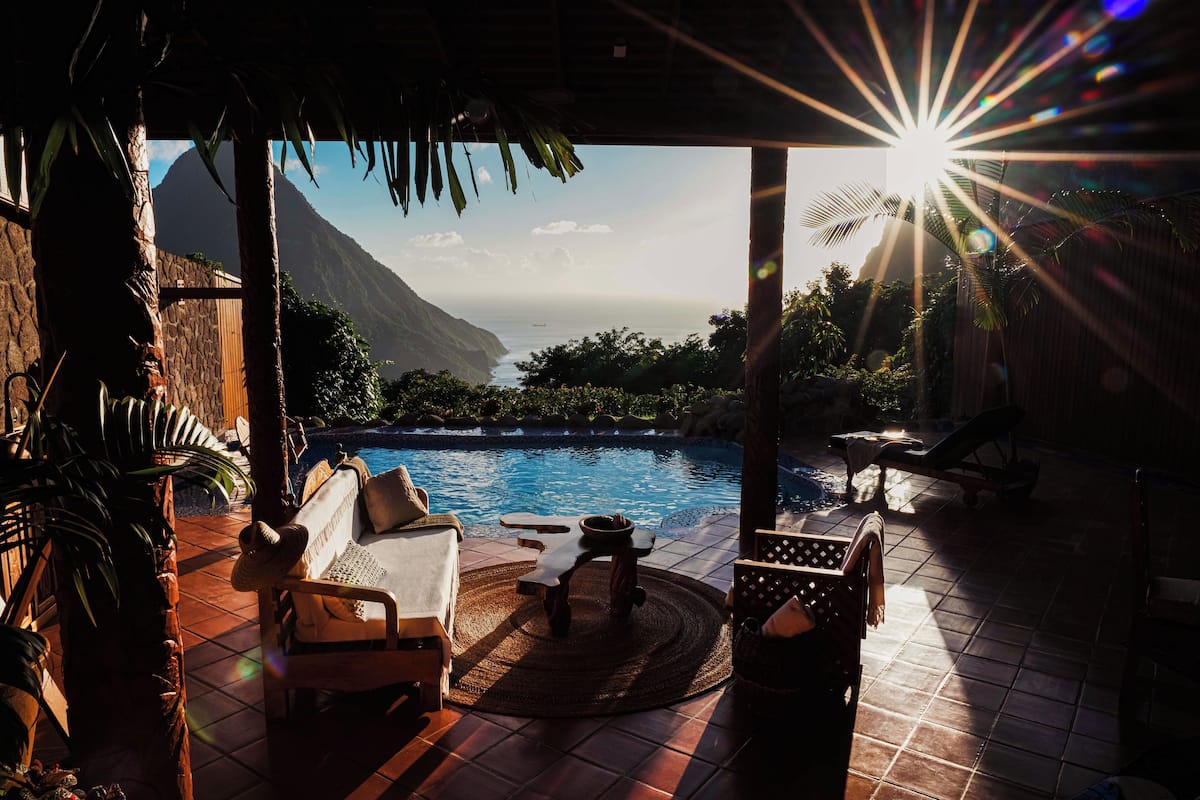 A poolside area with a view of a mountain and ocean, a lounge chair, a table, and a tiki bar.