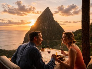 Una pareja cenando en una mesa con vistas a una montaña y al océano durante la puesta de sol.