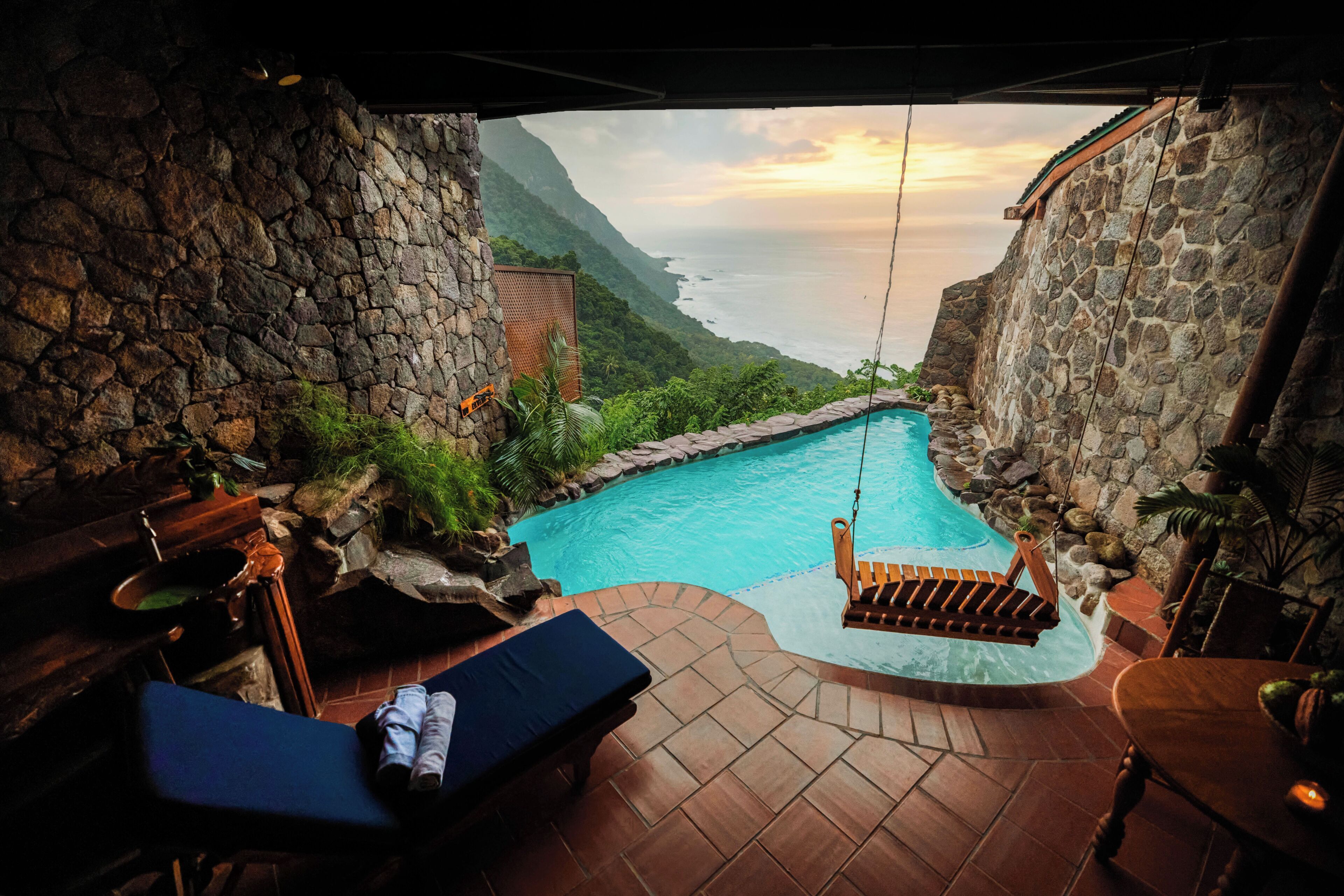 Piscina infinita de pedra com espreguiçadeiras, cadeiras de estar e uma mesa com velas, além de uma cadeira balanço de madeira.