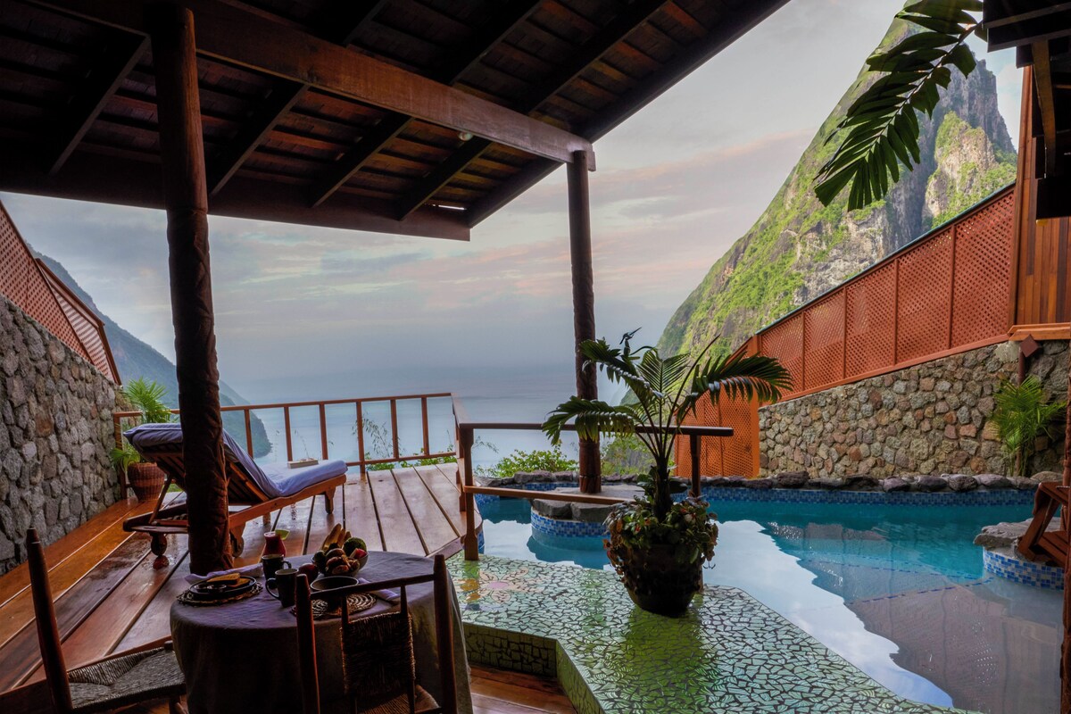 A pool area with a view of a mountain, wooden deck, lounge chairs, and a stone wall.