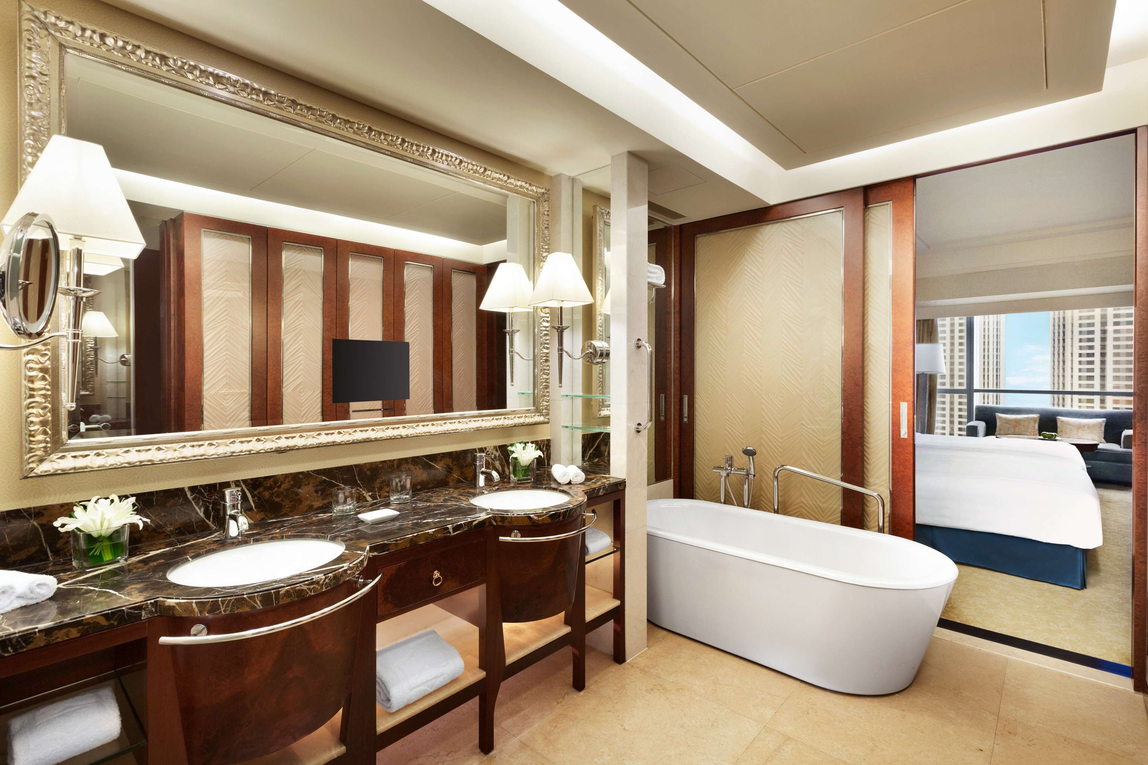 A luxurious bathroom with a large mirror, two sinks, a bathtub, and a view of a bedroom with a bed and a cityscape.
