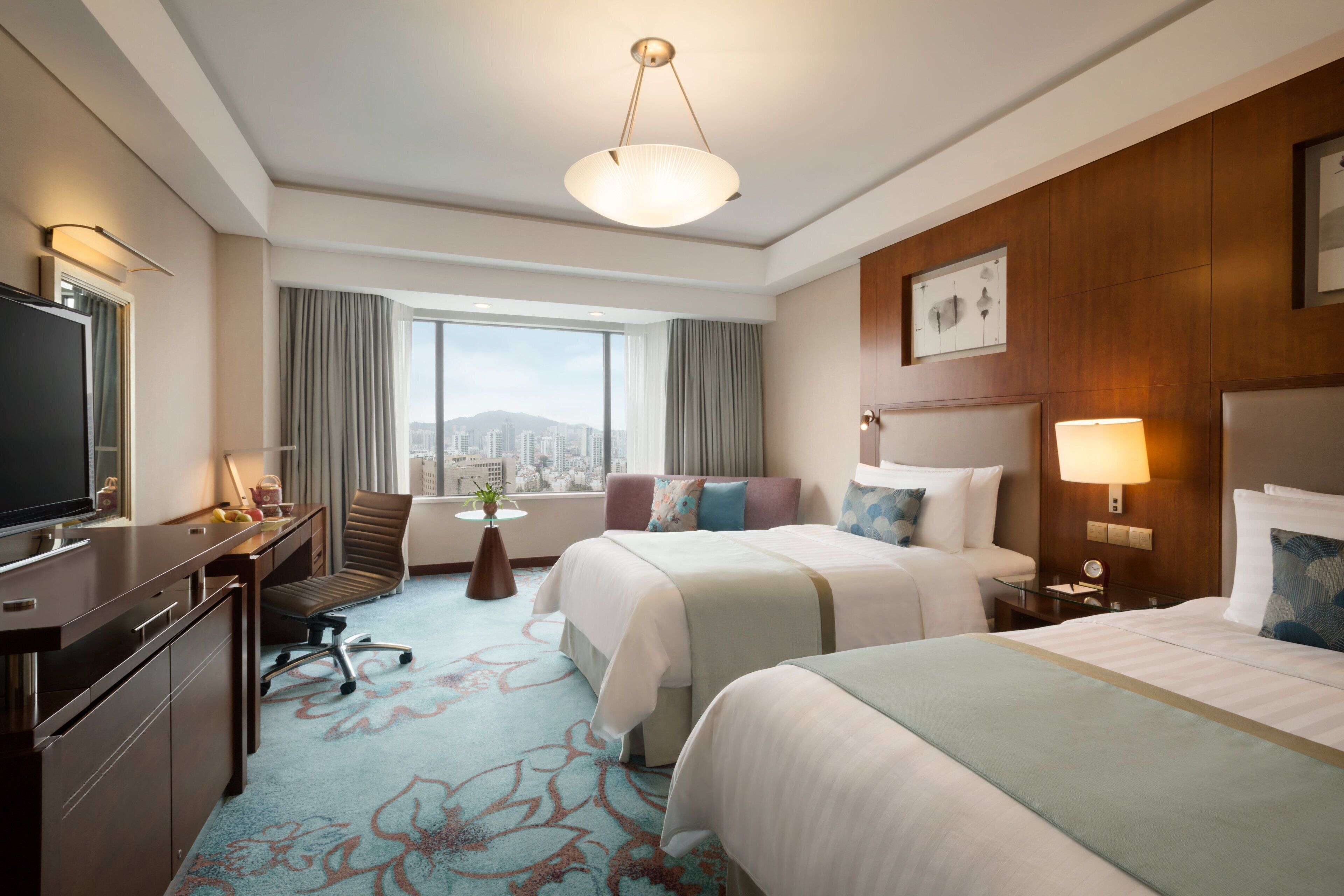 A hotel room with a large bed, a smaller bed, a desk with a TV, a chair, a lamp, and a view of the city.
