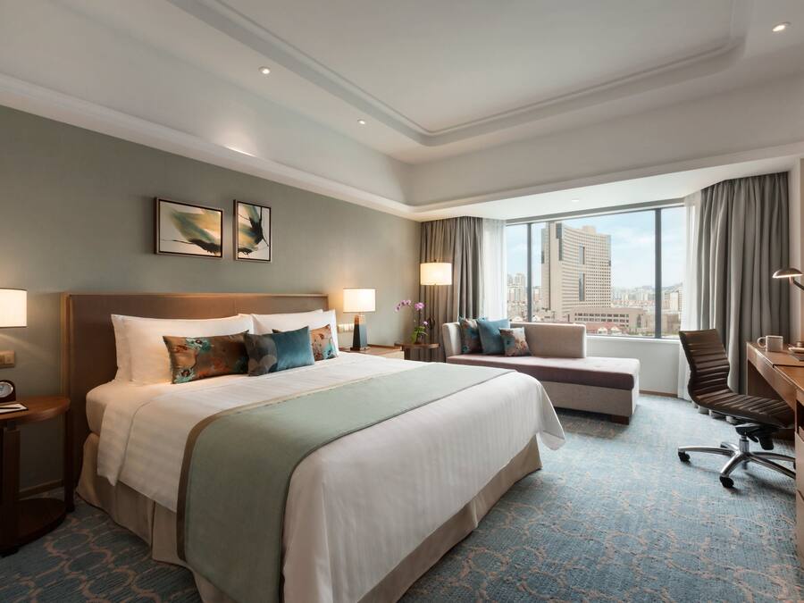 A hotel room with a large bed, a desk, a chair, and a sofa. There are two windows with curtains, a view of the city, and a clock on the bedside table.