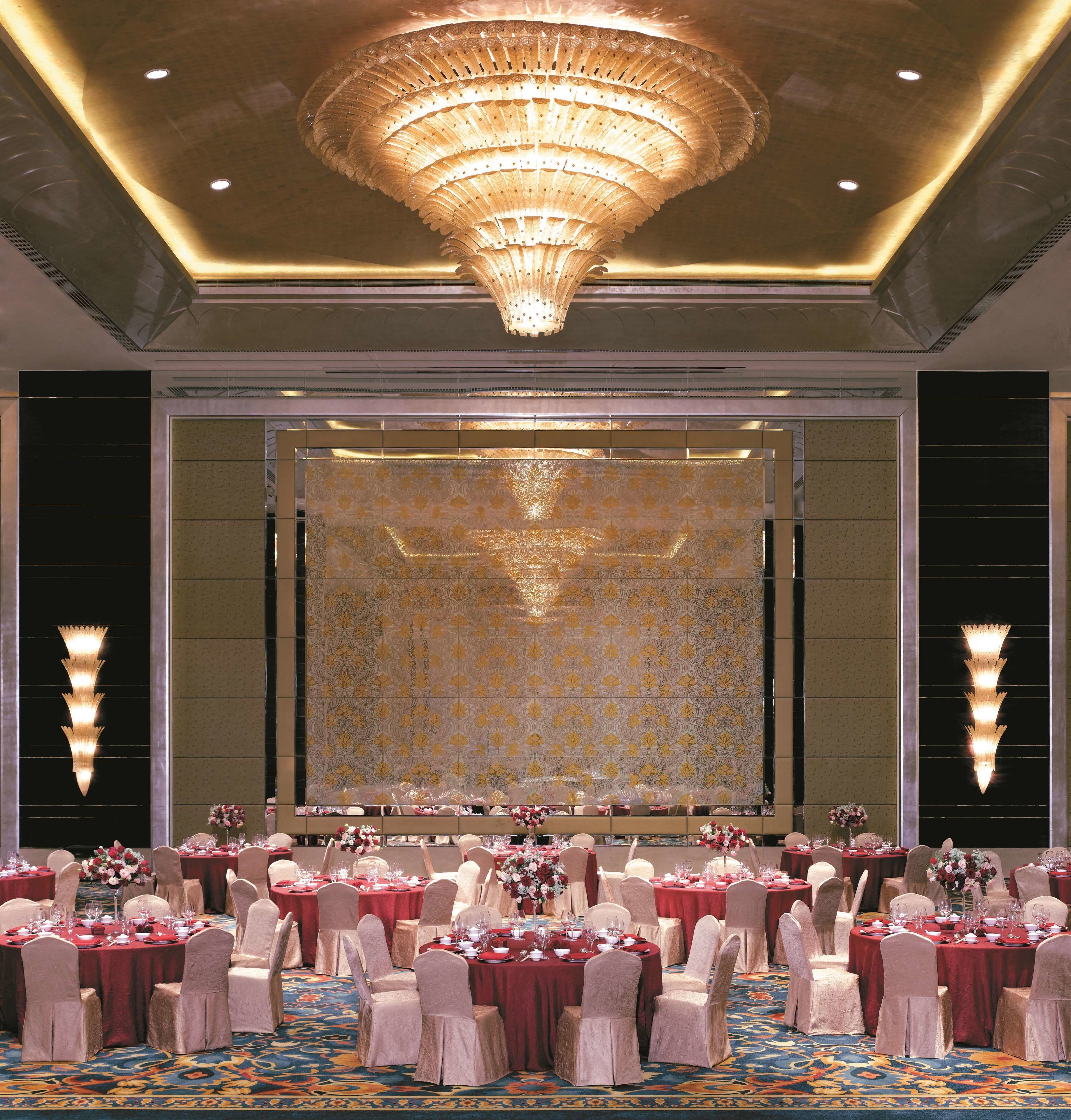 Salão de festas com mesas redondas preparadas para um evento formal, com um grande candelabro e papel de parede estampado.