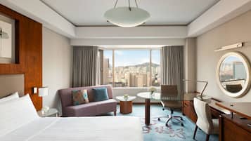 A hotel room with a large bed, a sofa, a desk, a chair, a round mirror, and a view of the city.