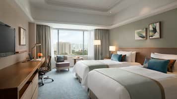 Habitación de hotel con dos camas, un escritorio con una computadora portátil, una silla, una mesita con una lámpara y un ventanal con vistas a la ciudad.