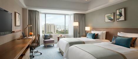A hotel room with two beds, a desk with a laptop, a chair, a small table with a lamp, and a large window offering a city view.