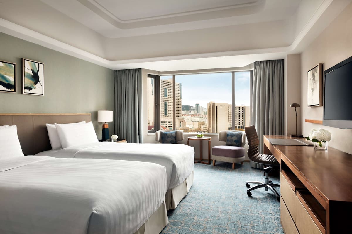 Ein Hotelzimmer mit zwei Betten, einem Schreibtisch, einem Stuhl, einem kleinen Tisch und Blick auf die Stadt.