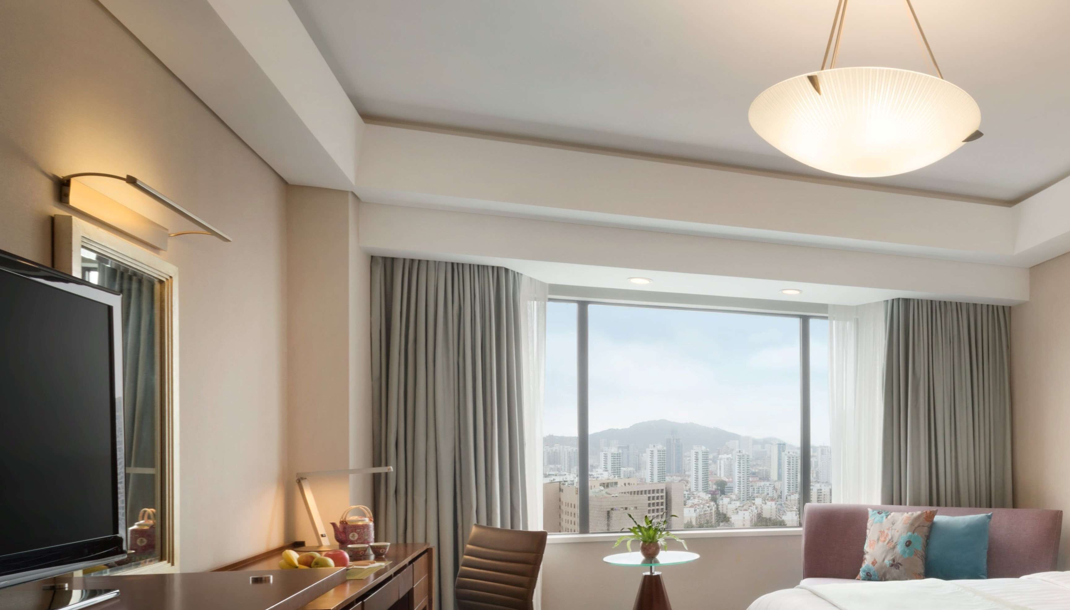 A hotel room with a large window offering a city view, a flat-screen TV, a dining table, and a comfortable bed.
