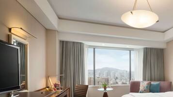 A hotel room with a large window offering a city view, a flat-screen TV, a dining table, and a comfortable bed.