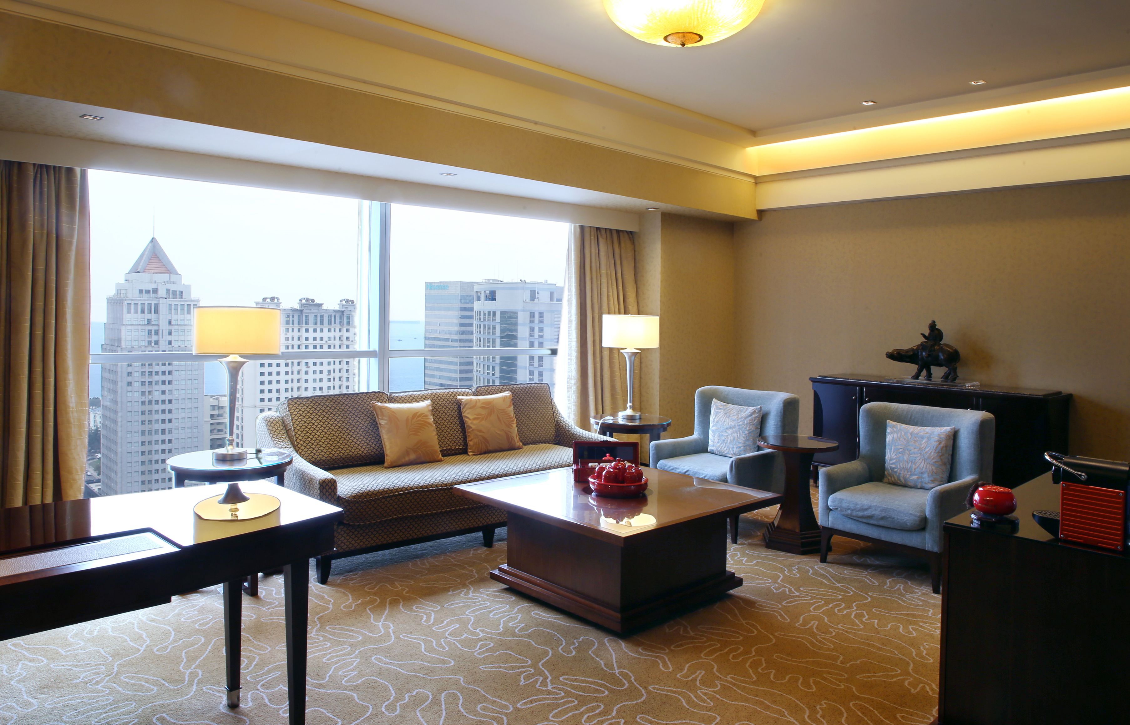 A hotel room with a sofa, armchairs, a coffee table, and a view of the cityscape through large windows.