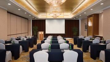 A conference room with rows of tables and chairs, a large screen at the front, and a chandelier.