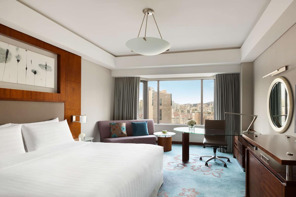 Ein Hotelzimmer mit einem großen Bett, einer Couch, einem Schreibtisch mit Stuhl, einem runden Tisch und Blick auf die Stadt durch große Fenster.