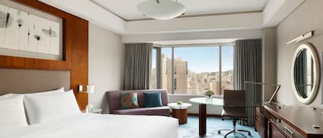 Una habitación de hotel con una cama grande, un sofá, un escritorio con una silla, una mesa redonda y vistas de la ciudad a través de amplias ventanas.