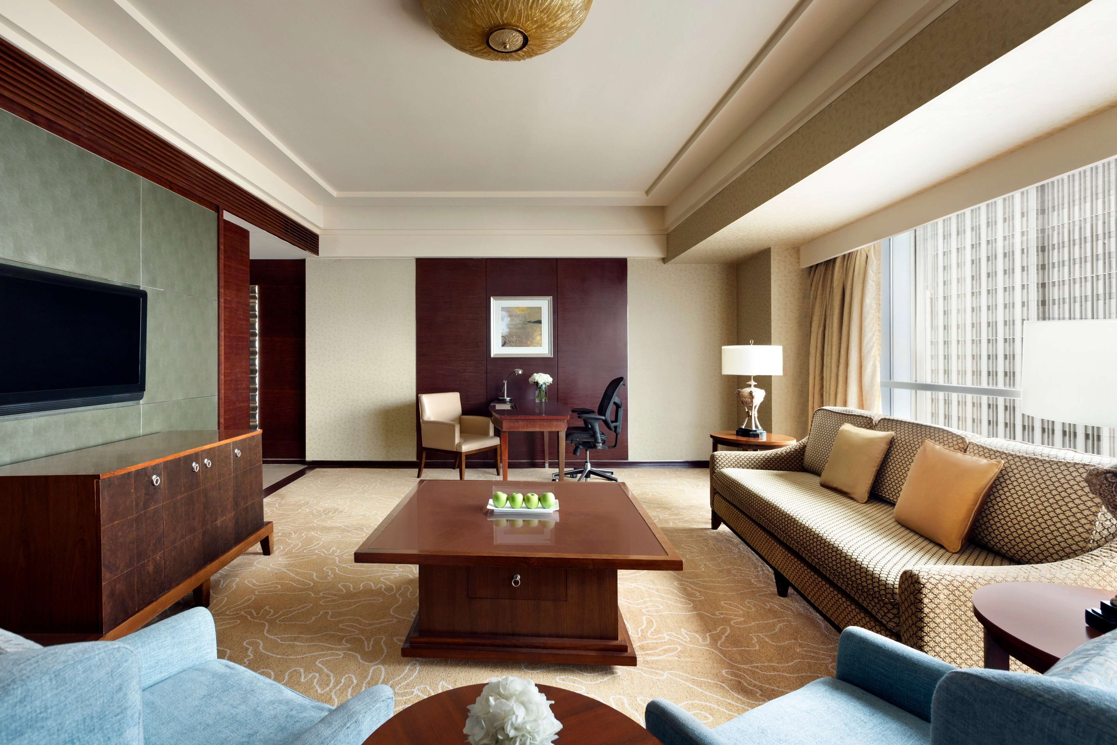 A hotel room with a large flat-screen TV, a wooden coffee table, a sofa, a chair, and a small table with a lamp.