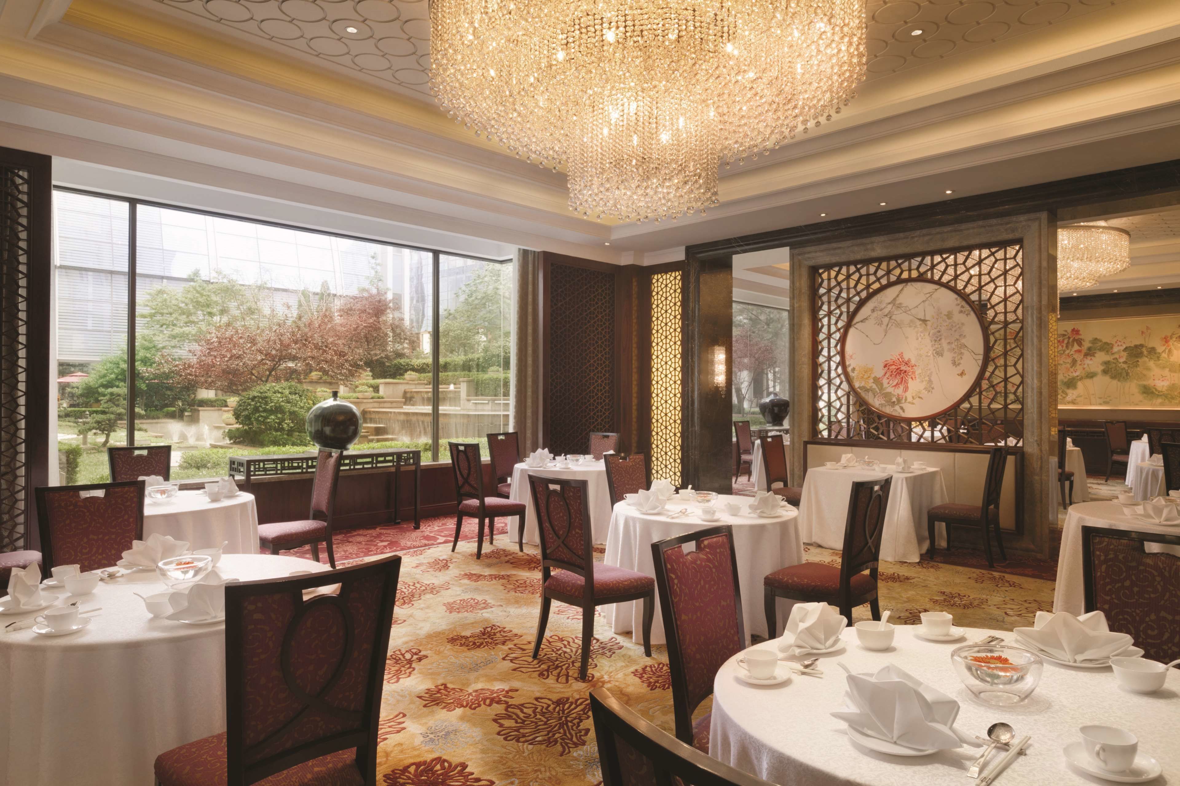 A dining area with round tables set for service, large windows offering a view of greenery, and a decorative ceiling light.