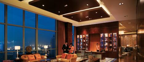 A spacious hotel lobby with a high ceiling, large windows offering a city view, and a bar area with shelves of bottles.