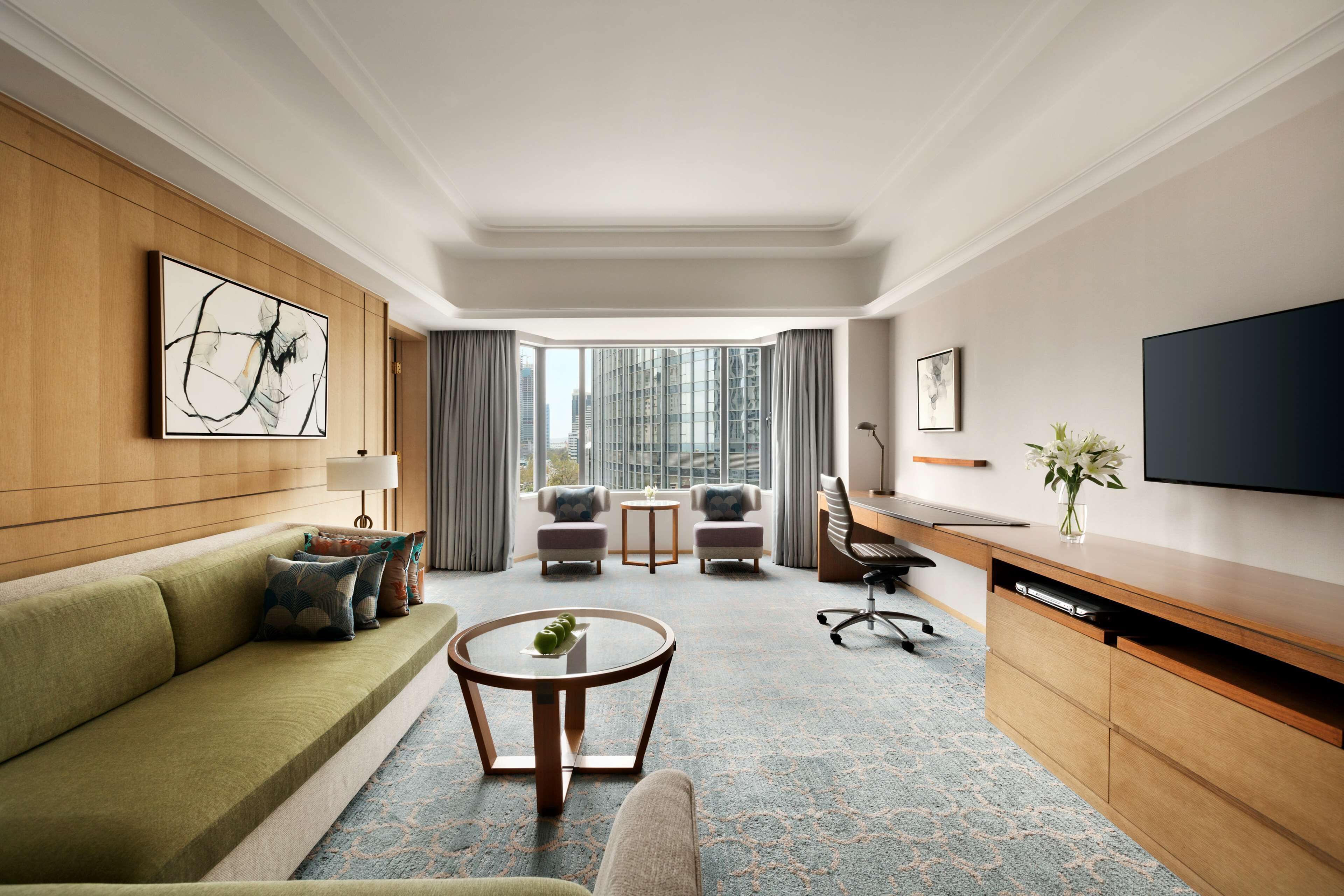 Een moderne hotelkamer met een groene bank, een ronde salontafel, een bureau met een flatscreen televisie, en uitzicht op de stad door grote ramen.
