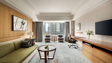 Een moderne hotelkamer met een groene bank, een ronde salontafel, een bureau met een flatscreen televisie, en uitzicht op de stad door grote ramen.