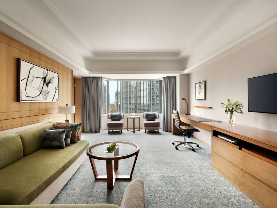 Een moderne hotelkamer met een groene bank, een ronde salontafel, een bureau met een flatscreen televisie, en uitzicht op de stad door grote ramen.