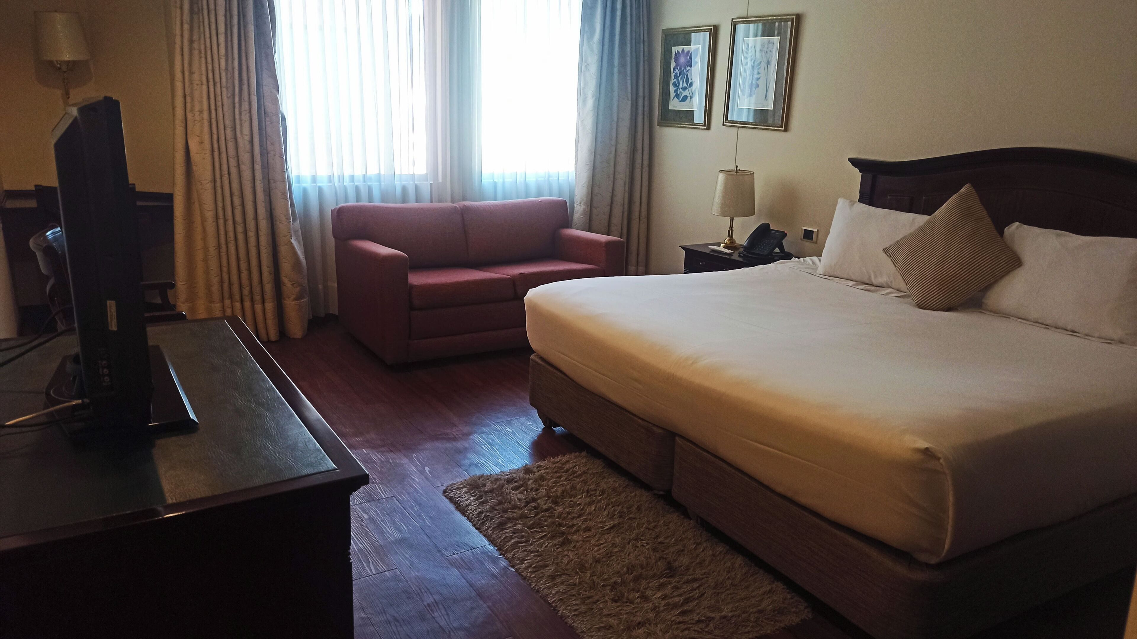 Une chambre d’hôtel avec un lit, un bureau avec un ordinateur, un fauteuil rouge, une table de chevet avec une lampe et deux tableaux encadrés au mur.