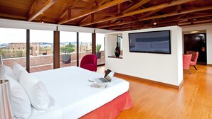 Una camera d'albergo moderna con un letto grande, una scrivania con un portatile, una TV a parete e vista sulla città attraverso ampie finestre.