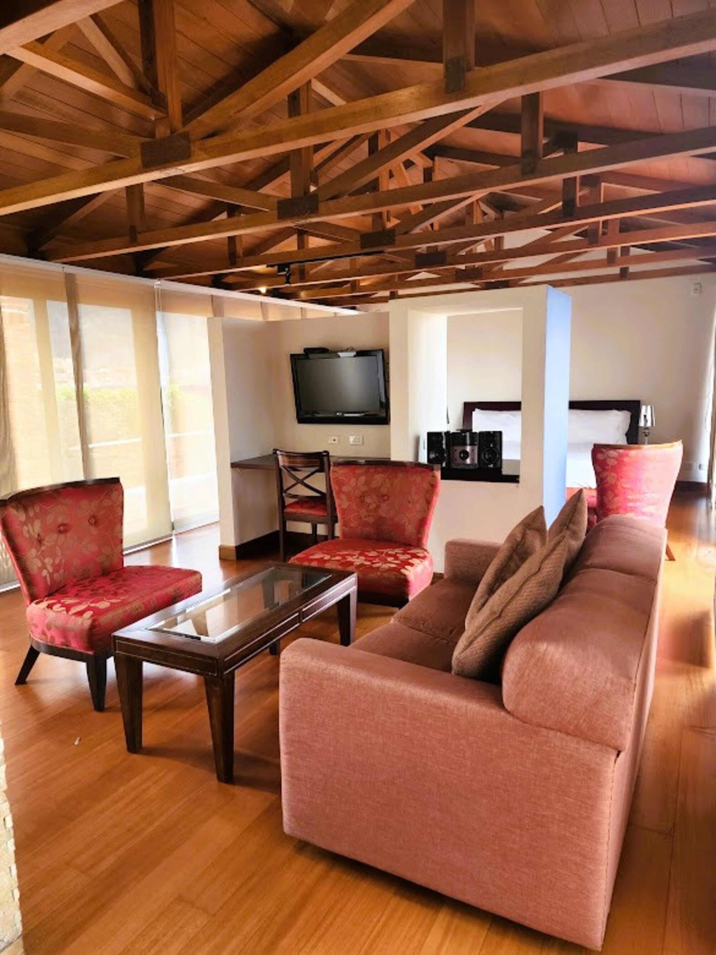 Una sala de estar amplia con techo alto, vigas de madera, un televisor de pantalla plana grande, un sofá, dos sillones y una mesa de centro.