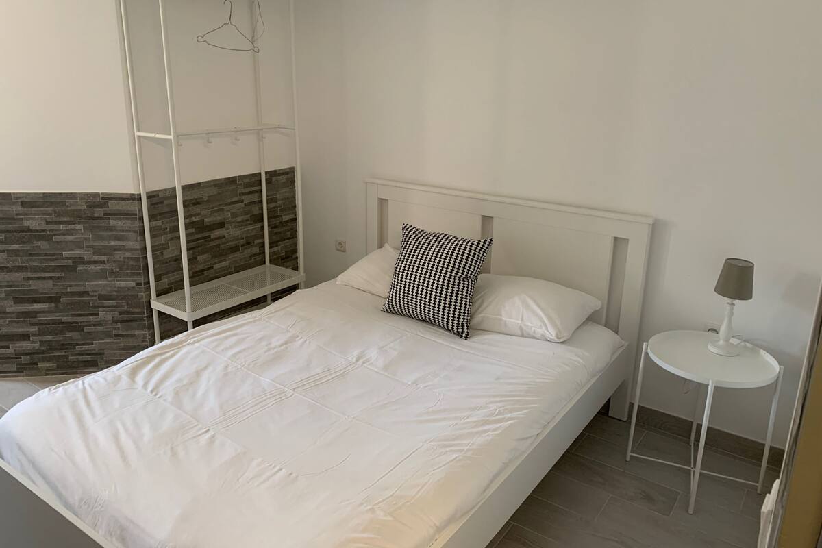 A neatly made bed with white bedding and a black and white patterned pillow, a small bedside table with a lamp, and a wall-mounted shelf with a clothes hanger.