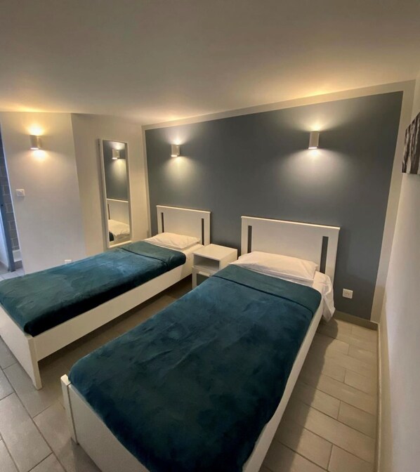 Habitación de hotel con dos camas individuales, cada una con cabecera blanca y colcha color turquesa. Hay mesitas de noche con lámparas, un espejo grande en la pared y una lámpara sobre las camas.
