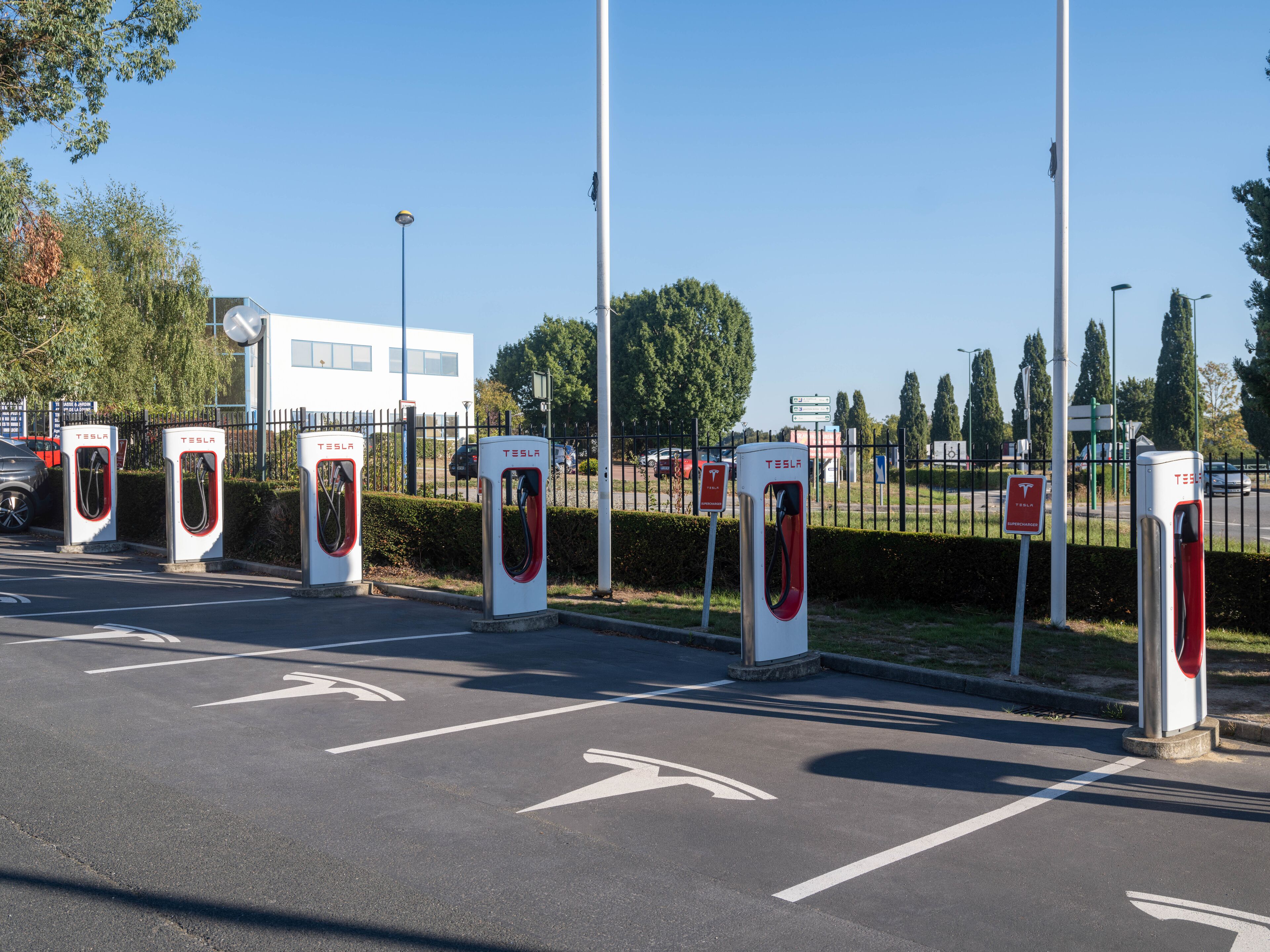 A parking area with multiple Tesla electric vehicle charging stations, a white building in the background, and directional arrows on the pavement.