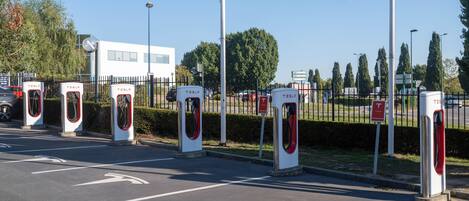 A parking area with multiple Tesla electric vehicle charging stations, a white building in the background, and directional arrows on the pavement.