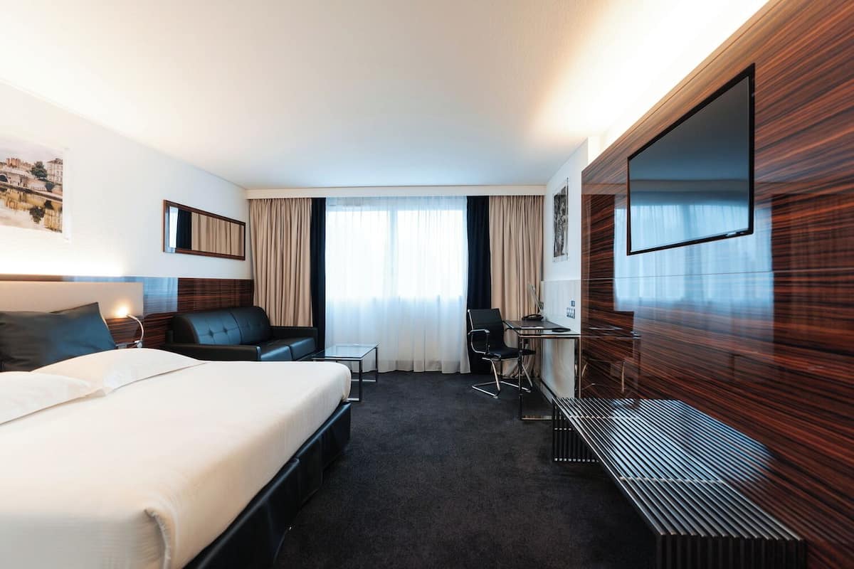 A hotel room with a bed, a desk, a chair, a sofa, and a large TV.