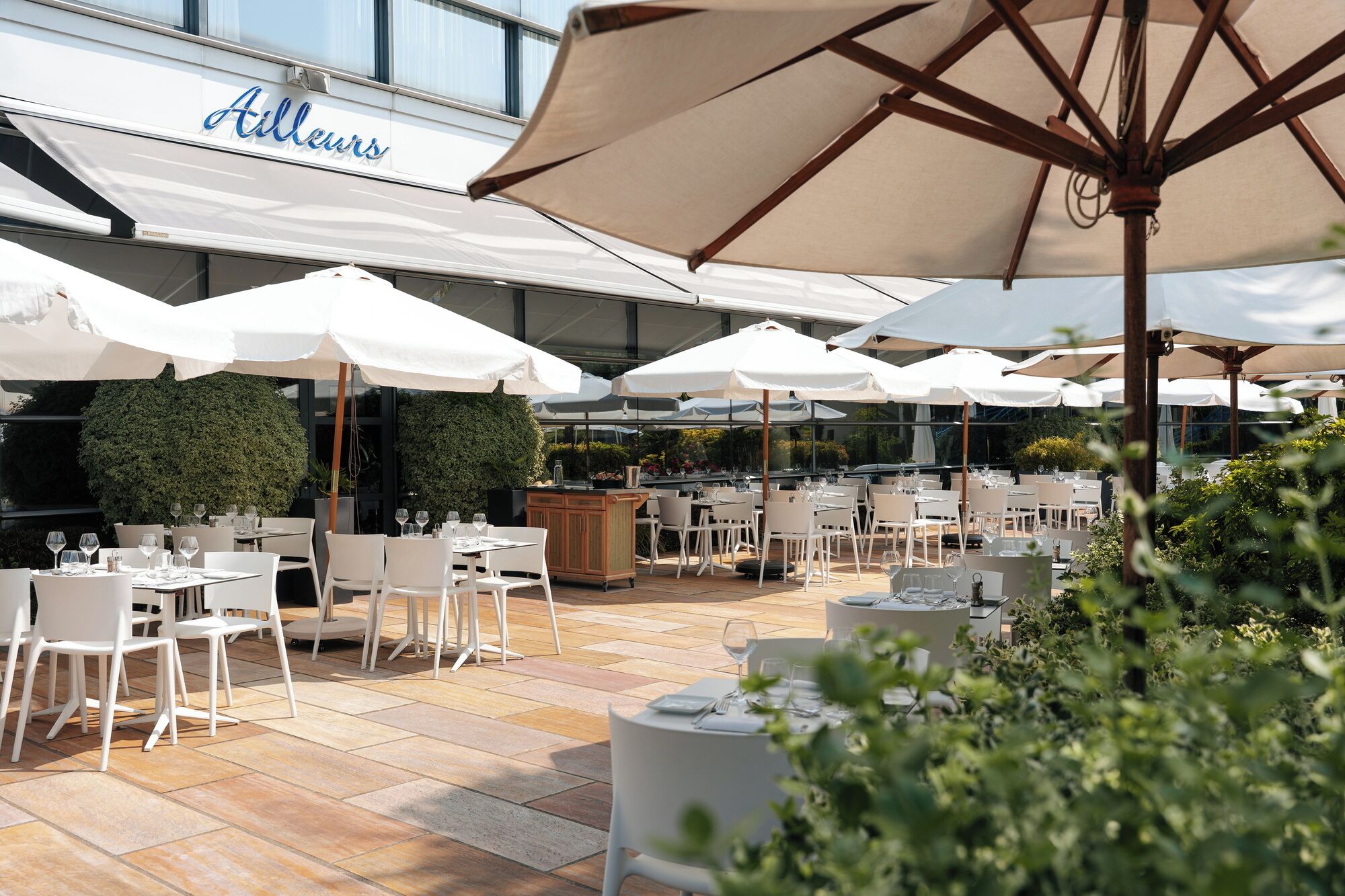 Un espace de restauration en plein air avec des tables et des chaises blanches, de grands parasols et une enseigne indiquant « Allleurs ».
