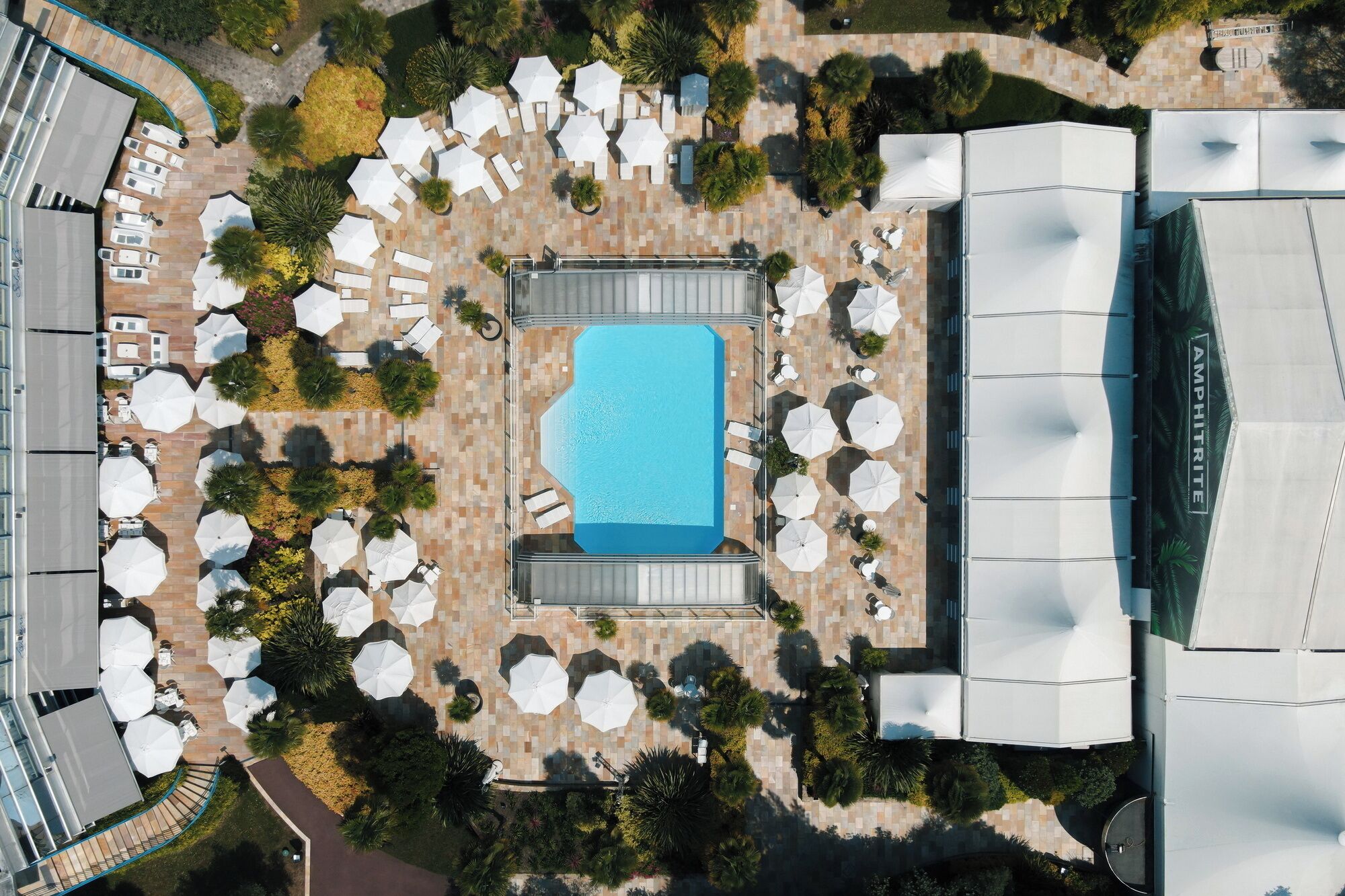 An aerial view of a resort with a central swimming pool, surrounded by sun loungers and umbrellas, and adjacent to a large amphitheater.