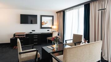 Habitación de hotel con televisión de pantalla plana, escritorio con lámpara, silla, mesa con un jarrón de flores y cama con maleta.