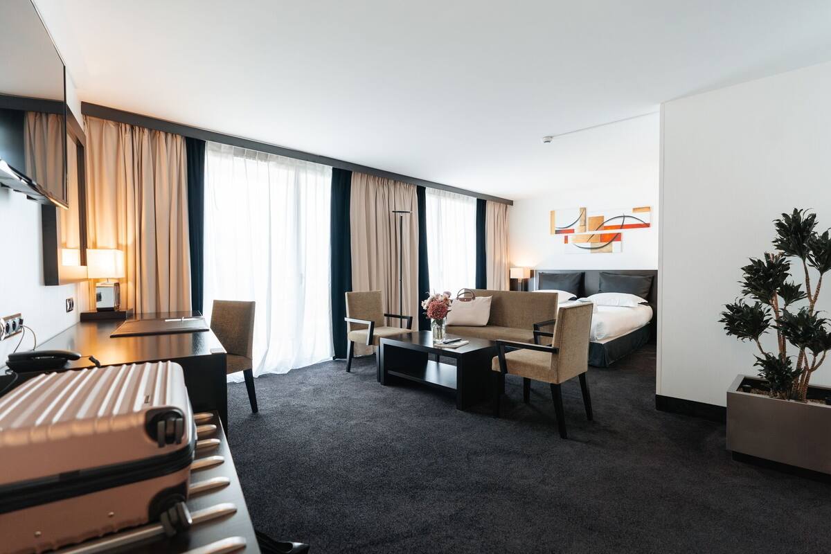 A hotel room with a bed, a sofa, a desk, and a coffee table. There is a plant in the corner and a window with curtains.
