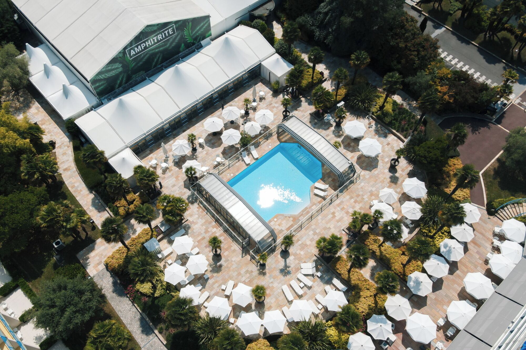 Vista aérea de un hotel con piscina rodeada de tumbonas y sombrillas, una zona cubierta con una carpa y un jardín ajardinado.