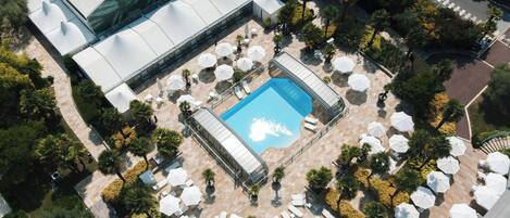 An aerial view of a hotel with a swimming pool surrounded by sun loungers and umbrellas, a covered area with a tent, and a landscaped garden.