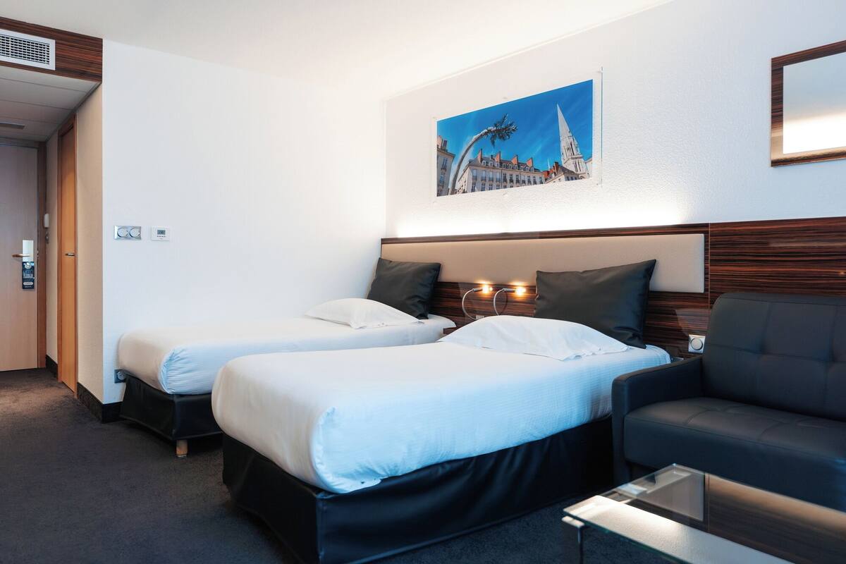 A hotel room with two beds, a sofa, a glass coffee table, and a painting of a building on the wall.