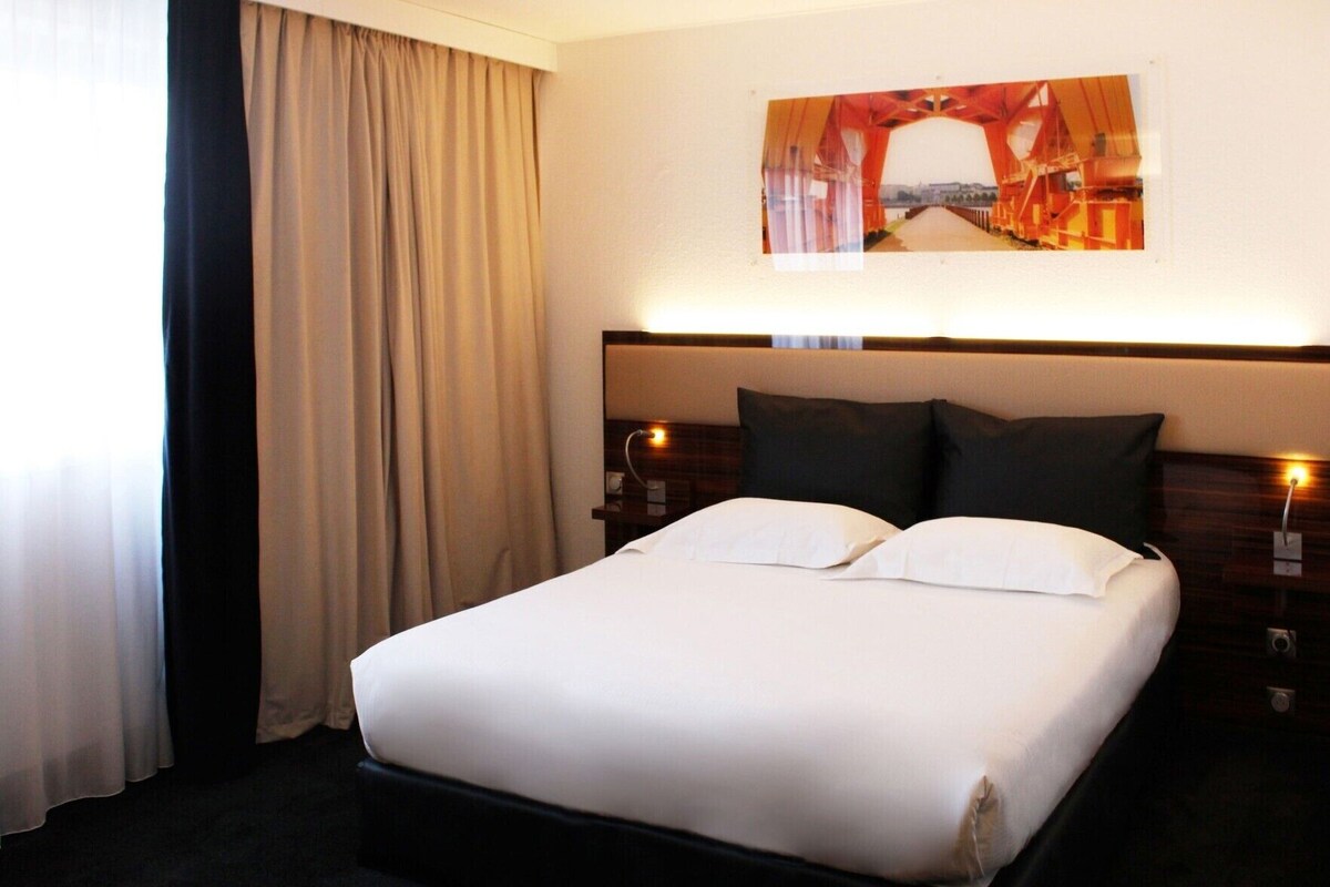 Une chambre d’hôtel comprenant un lit, deux tables de chevet, une tête de lit, une fenêtre avec des rideaux et un tableau accroché au mur.