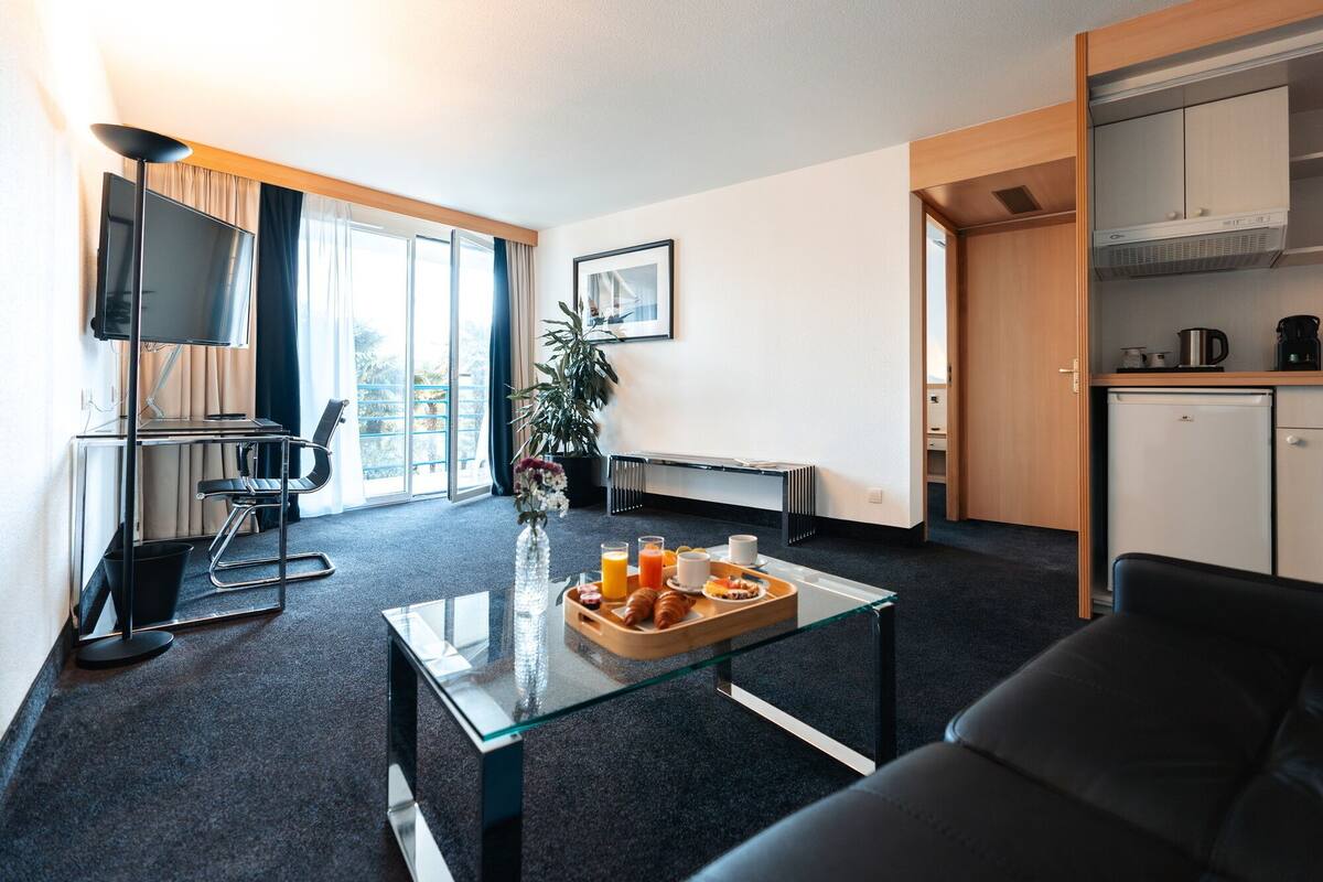 A modern hotel room with a glass coffee table, a black leather sofa, a desk with a chair, a television, and a kitchenette.