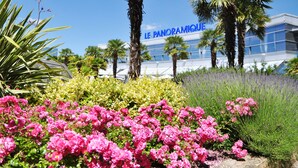 Un bâtiment moderne, sur lequel on peut lire l’inscription « LE PANORAMIQUE », entouré d’une végétation luxuriante et de fleurs roses.