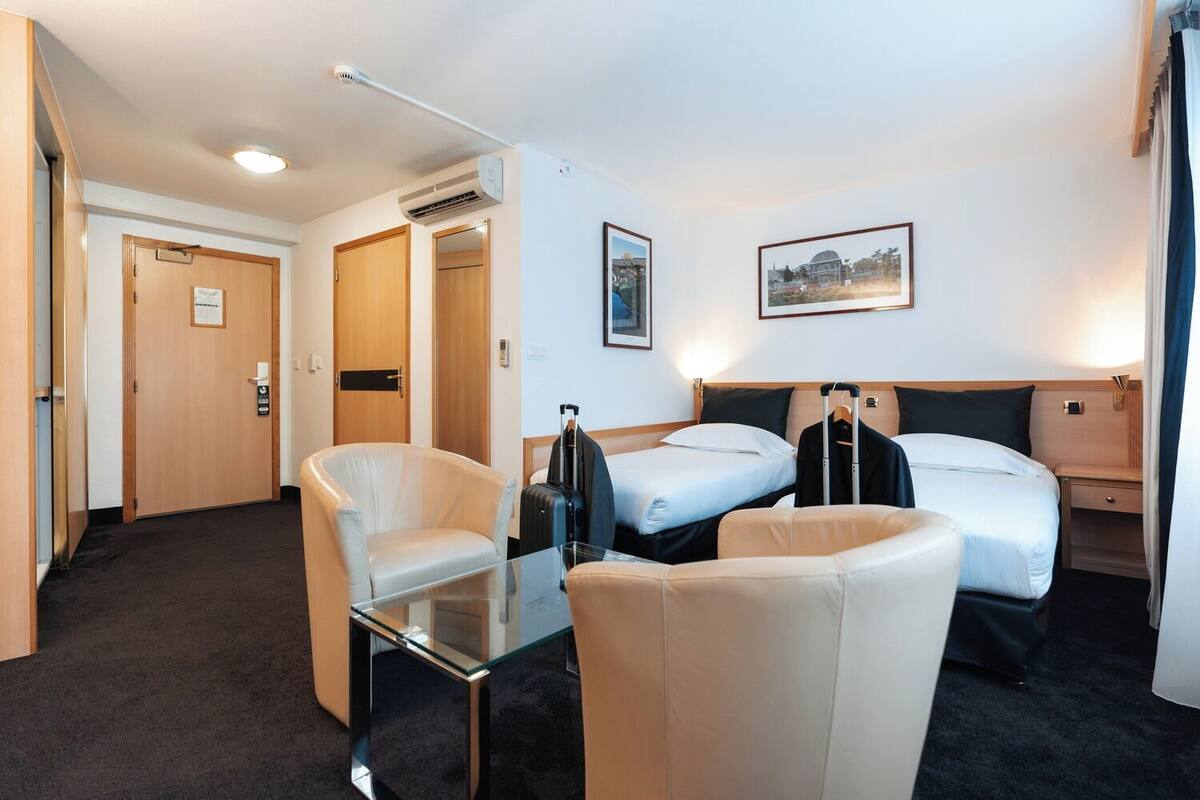 A hotel room with two beds, a glass table, a chair, a coat rack, and a framed picture on the wall.